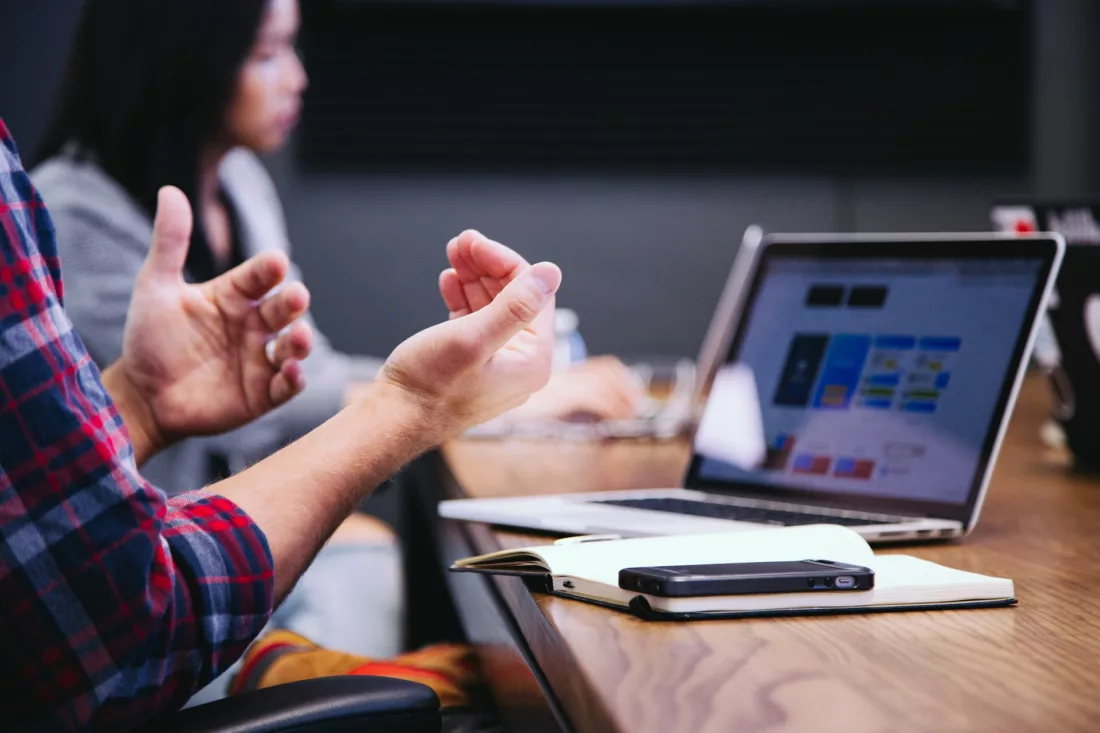 A close-up of a person's hands gesturing during a meeting, with a laptop displaying a colorful interface and a notebook beside it on a wooden table. Another person is blurred in the background, contributing to a discussion about 社群操作.