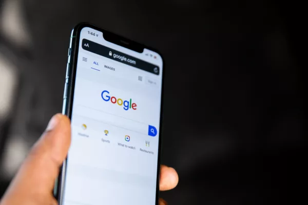 A hand holds a smartphone displaying the Google homepage. The screen shows Google’s logo and search bar, along with icons for search options below it. The background appears blurred, emphasizing the mobile device and its content.